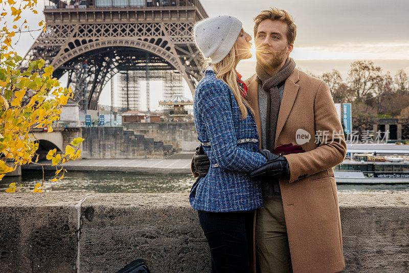 女友在巴黎埃菲尔铁塔前亲吻男友，傍晚，阳光在她的双唇和男友的脸颊之间