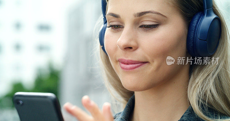 特写的女人听音乐与耳机和电话。女性脸上戴着无线蓝牙耳机。一位女士站在城外用智能手机选歌
