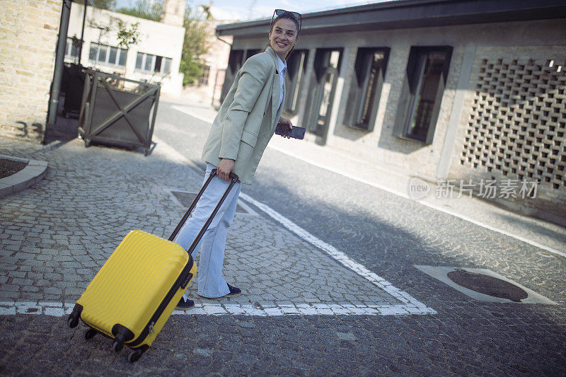 一个面带微笑的女人提着一个黄色的手提箱等待运输。她很高兴终于要去度假了。