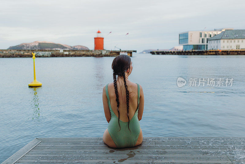 女性在泳装坐在码头上看红色灯塔在Norwa的后视图