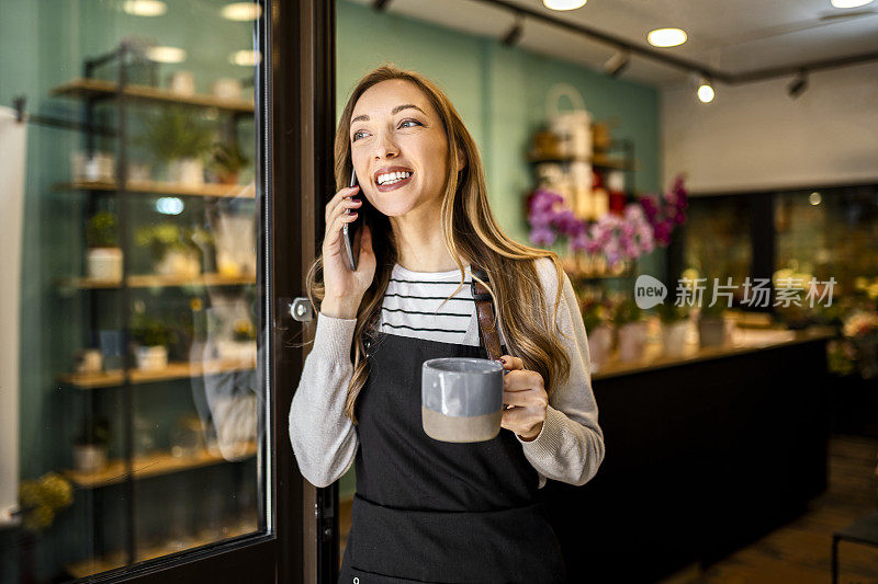 快乐的年轻花匠在花店用智能手机拿着一杯咖啡
