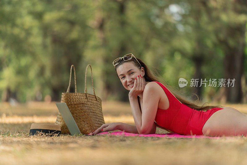 美丽的白人女性性感的女人在红色泳装比基尼戴着太阳镜躺在地上工作在她的电脑笔记本电脑放松在树林里的海滩