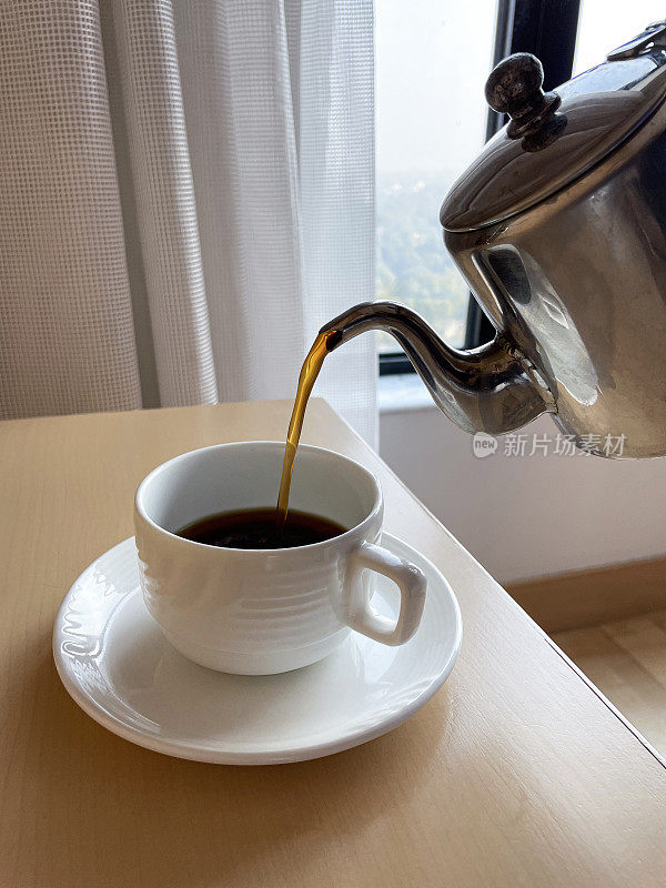 特写图像新鲜的黑咖啡从不锈钢咖啡壶的壶嘴，白色的杯子和碟子，酒店客房服务，重点在前景