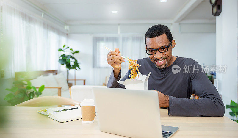 非洲商人在办公室享受中餐的肖像午休在线会议，员工拿着筷子吃外卖面盒餐虚拟在家办公室的工作场所。