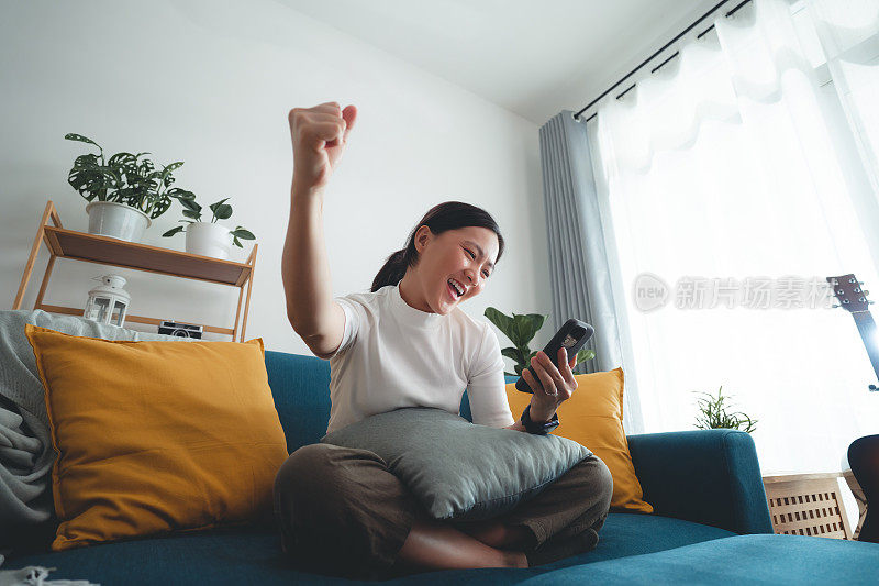 亚洲女人坐在家里的客厅里，用智能手机开心兴奋地做了一个获胜的手势。