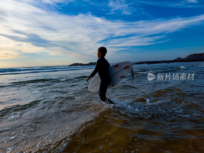 一个年轻而健壮的男孩在日落时分穿过海滩上的水去冲浪