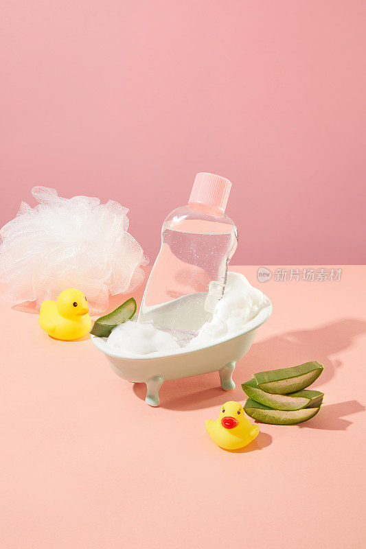 瓷浴缸内装有一瓶没有标签的婴儿洗漱用品，透明液体填充，粉红色背景上装饰有vera、沐浴海绵和橡皮鸭。拷贝空间，前视图