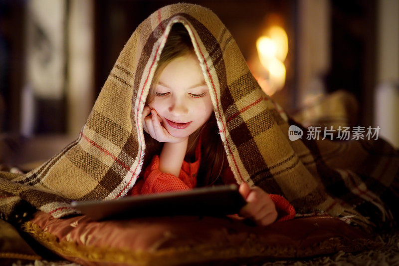 可爱的小女孩在圣诞节晚上壁炉旁使用平板电脑