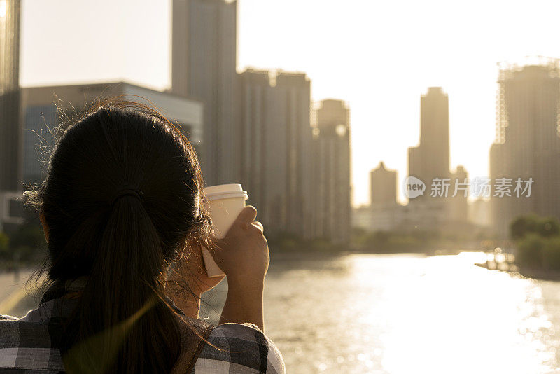 拿着咖啡杯的女孩，欣赏着夕阳下的城市天际线