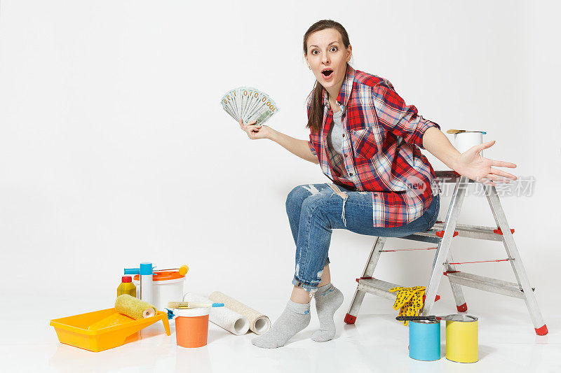 一个女人手里拿着一捆美元，现金，坐在梯子上，手里拿着装修公寓用的工具，孤立在白色背景上。壁纸配件用于粘漆工具。修理家里的概念。