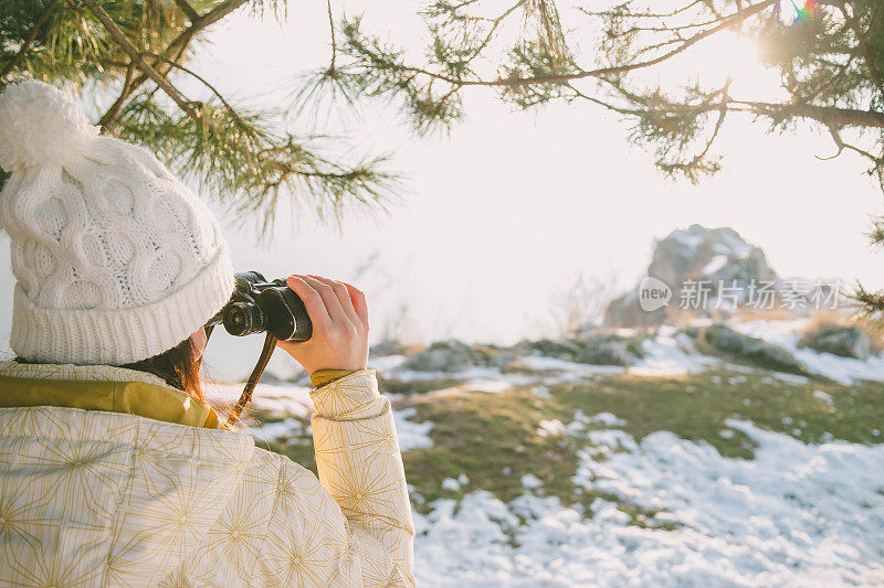 冬天的森林里，戴着白帽、背着背包的女孩用双筒望远镜看东西