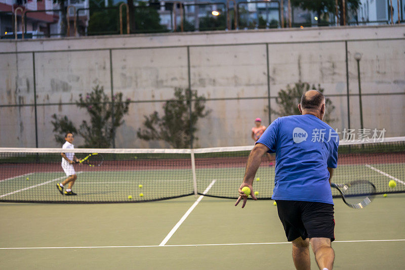 教练正用正手向孩子们打网球，选手在硬地场上专注于前景，教练手上有签名