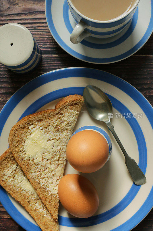 蓝色和白色康沃尔陶器的图像，茶盘和鸡蛋杯与煮鸡蛋，茶匙，全麦黄油面包片，盐瓶和杯和碟子含有奶茶，木纹背景，升高的视野