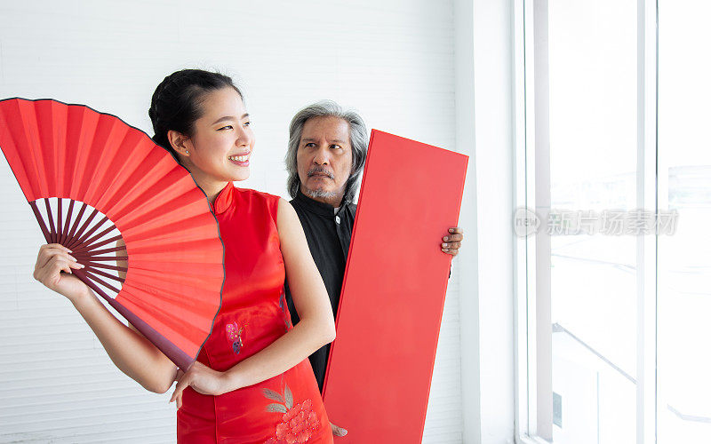 选择聚焦美丽快乐的亚洲富婆穿着红色旗袍或连衣裙，微笑着，拿着折扇与模糊的男性保镖，庆祝中国新年。宗教与节日观念。
