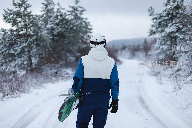 年轻的男性滑雪板手拿着滑雪板，走在被雪树环绕的小路上