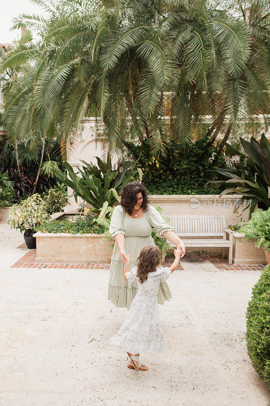 在佛罗里达州棕榈滩的一个托斯卡纳风格的花园里，一个古巴裔美国4岁小女孩和她31岁的古巴妈妈跳舞的瞬间，她都有棕色的卷发，穿着鼠尾草绿和白色的春装和白色的皮凉鞋