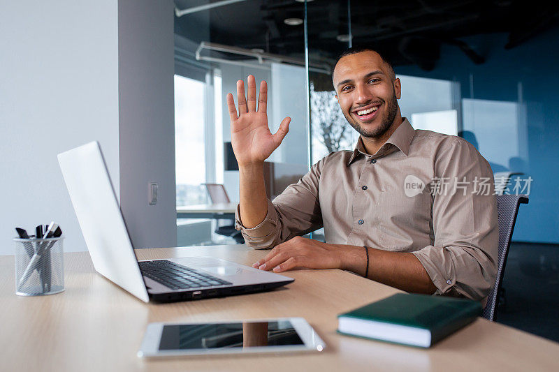 满意的非洲裔美国商人在办公室，在笔记本电脑上工作，敲击键盘，挥手打招呼，看着相机