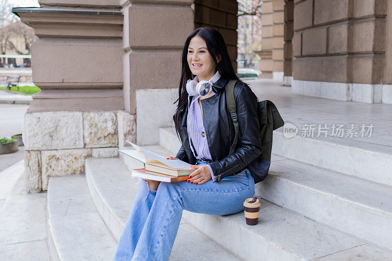 年轻漂亮的女大学生背着双肩包坐在大学楼梯上。她戴着耳机，穿着皮夹克，等待着大学课程的开始。