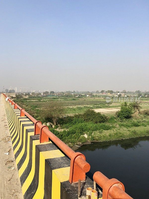 这是印度德里的公路桥，用于在河流上行驶，路边的混凝土墙上有黄色和黑色的危险条纹，铁安全栏杆漆成亮红色的橙色，绿色的农田，河边的农场和庄稼