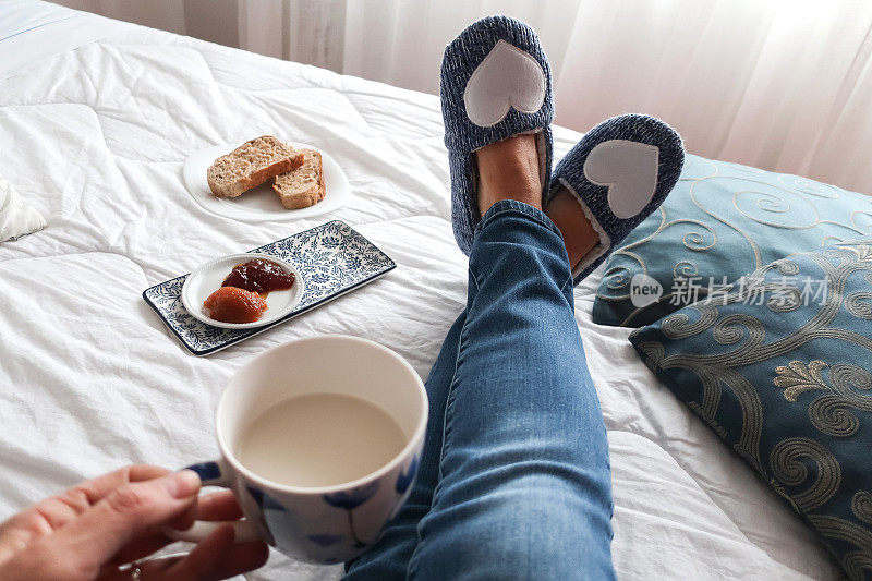 个人观点的高级女人腿放松在床上与蓝色拖鞋在下午茶休息