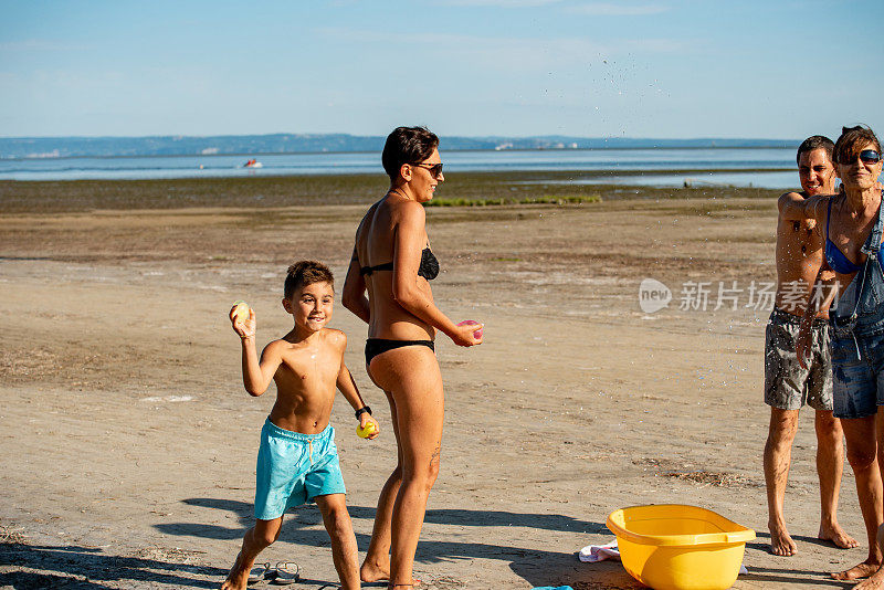 快乐的家庭站在海滩上，顽皮的儿子玩水炸弹-库存照片
