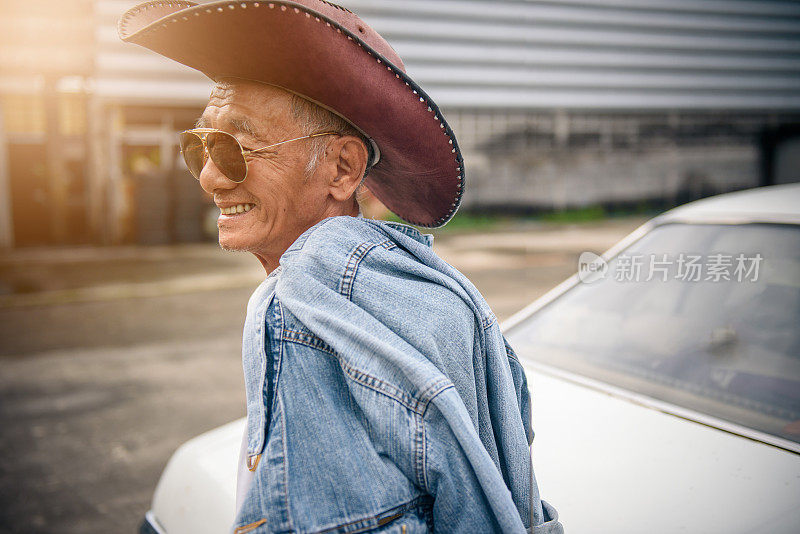 年长的亚洲男人在牛仔夹克摆姿势与古董车