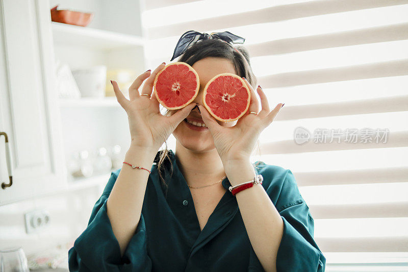 一个年轻女子拿着一个葡萄柚，捂着眼睛，坐在厨房里享受着休息日。一个快乐的年轻女子拿着一个葡萄柚，这是一种万能的美容工具