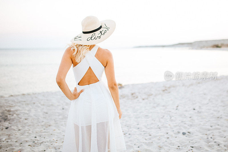 美丽的女人戴着草帽，穿着白色的衣服走在海边的海滩上