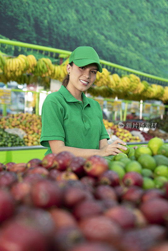 一位拉丁裔女子在杂货店里看着镜头，一边整理着她的水果和蔬菜