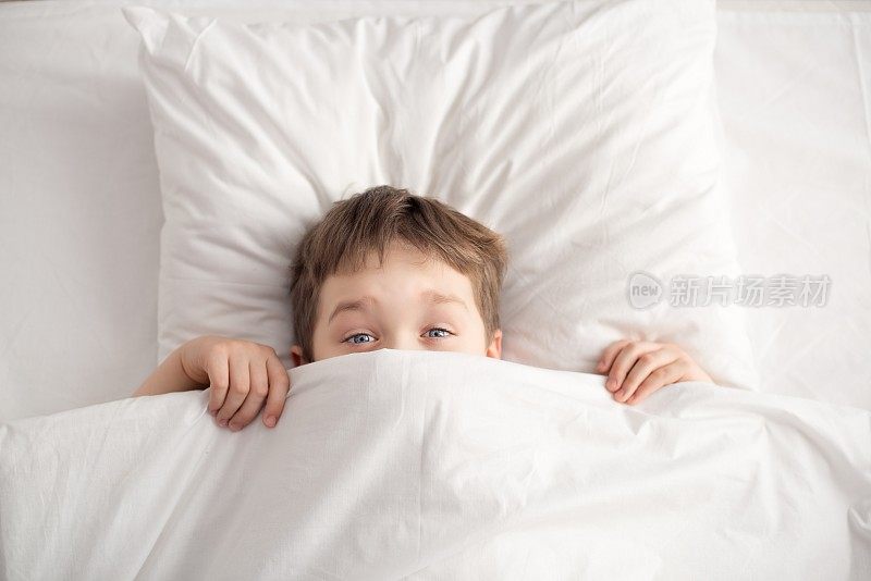 快乐的男孩躺在白色的床上，盖着白色的毯子