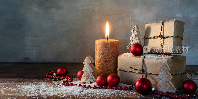 两个礼盒和一个燃烧的蜡烛，红色的圣诞装饰物和小木制玩具树在一些雪地上的乡村木材，复古的背景与大的复制空间，全景格式