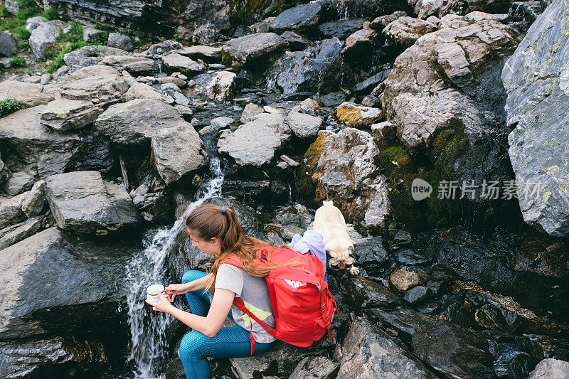 一个背着红色背包的女人和一只狗在徒步旅行后休息，并从挪威河打水