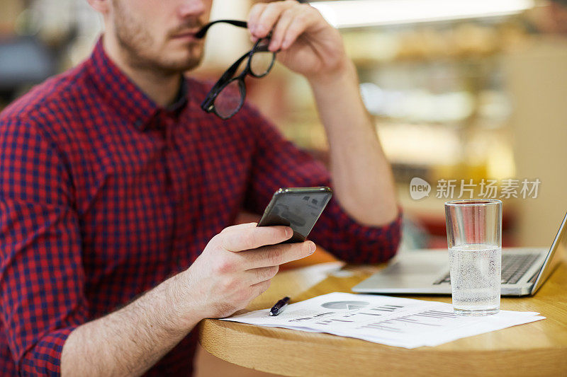 镜头的特写是一位沉思的、忙碌的分析师，他穿着休闲衬衫坐在桌子前，手里拿着财务报告、笔记本电脑和一杯水，一边咬着眼镜，一边使用智能手机，他在进行数据研究