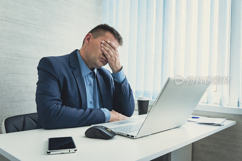 疲惫或精疲力竭的人闭着脸，双手坐在办公室的电脑前。商人用手遮住脸和眼睛。facepalm指