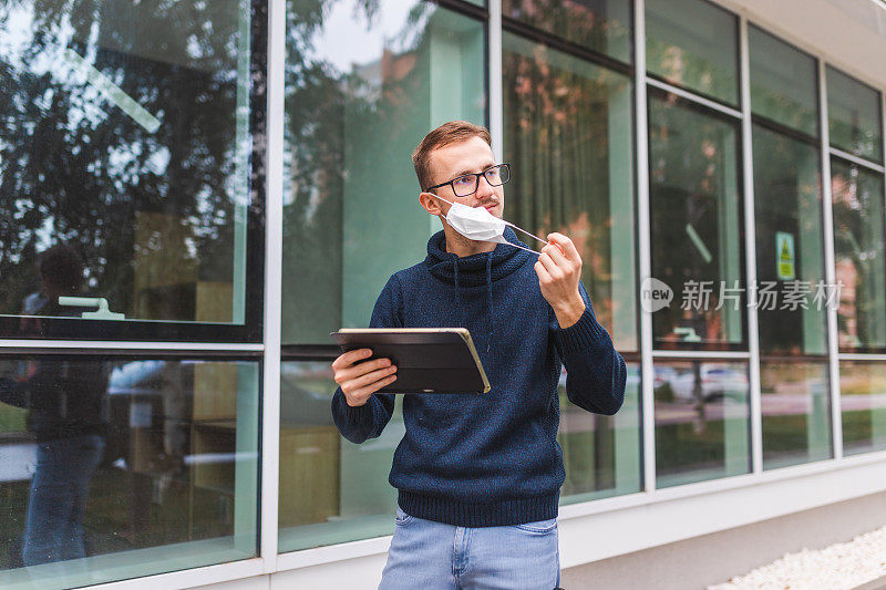 机场外，一名戴眼镜的年轻人脱下防护面罩，手持数码平板电脑