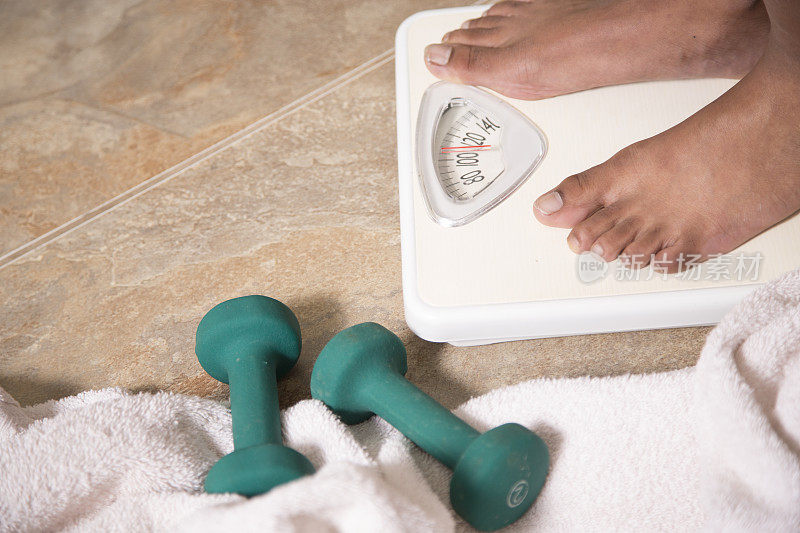 健康的生活方式-非裔美国妇女踩浴室磅秤。她的运动器械和浴巾就放在秤旁边的地板上。