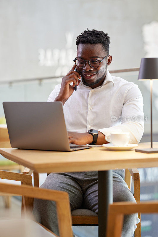 一位忙碌的非洲商人坐在自助餐厅里，一边用笔记本电脑打字，一边和客户打电话。