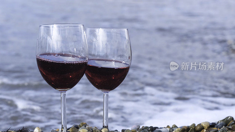 阳光明媚的沙滩上，喝着红酒，享受浪漫的一天。的概念。两个透明的酒杯和红酒放在卵石海滩上，以海浪和大海为背景。