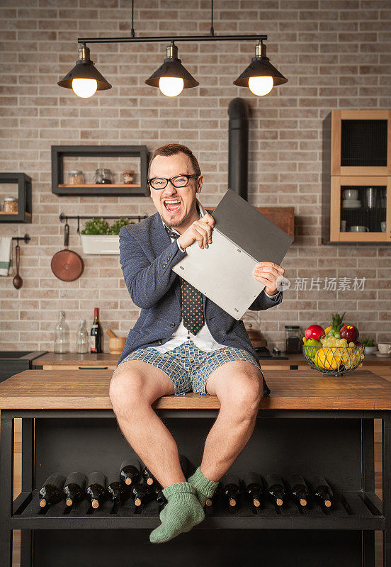 愤怒的帅哥穿着西装和内裤在家里上网，坐在厨房桌子上砸他的笔记本电脑