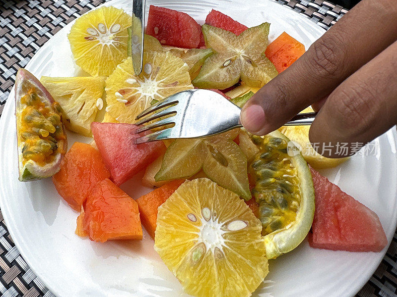 新鲜的热带水果沙拉，白色盘子里有切片的橙子、菠萝、杨桃、西瓜和西番莲，被一个认不出来的人用刀叉切开，桌面，高视图