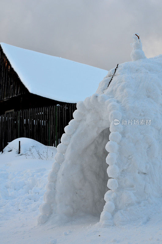 典型的冬季山地儿童娱乐。雪屋屋顶上的雪人。入口的边缘像蕾丝。压痕。有木栅栏和谷仓的田园牧歌