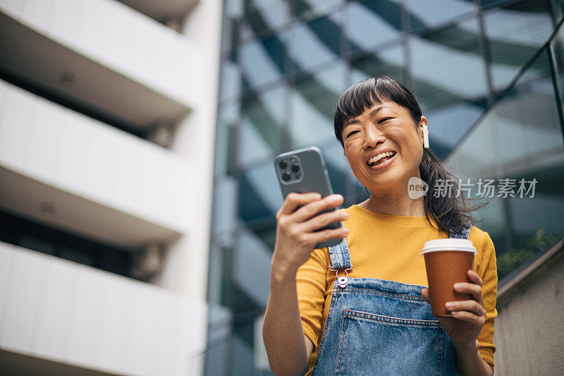 一名日本女子拿着智能手机和一杯咖啡，看着手机屏幕，背景是一栋办公楼