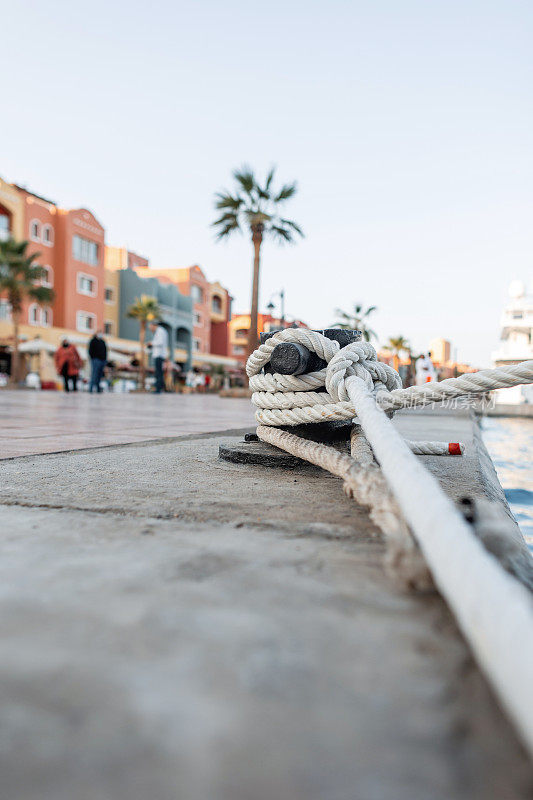 在一个有棕榈树的港口城市，一艘船被一根白色的绳子拴在一个金属系柱上。游艇停在码头上，特写。在海上休息和旅行