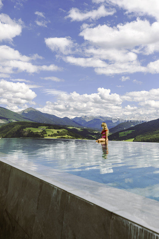 一个穿着红色泳装的年轻女子站在无边泳池边，享受着美景，在阳光明媚的夏日，意大利的多洛米蒂，泳池边的自然景观一览无余