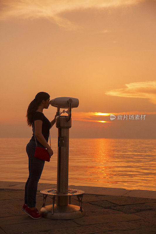 年轻女子透过投币双筒望远镜眺望海洋漫步海上日落