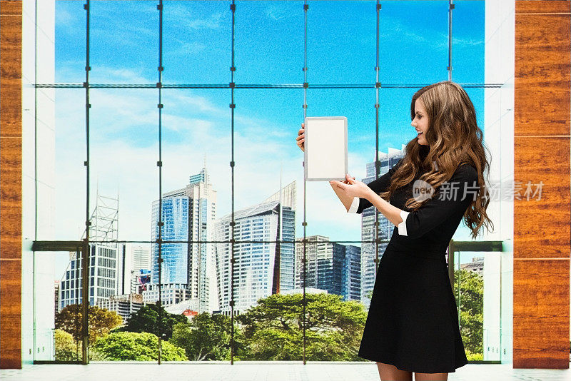 一位女商人站在办公室大厅里，手里拿着一台平板电脑，正在展示屏幕