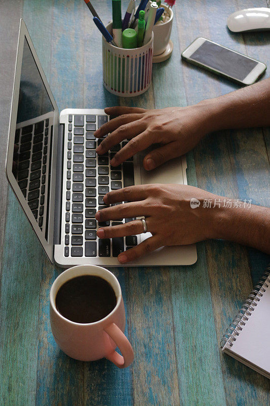 一个不知名的人在办公室工作站的笔记本电脑上打字，周围是咖啡杯，黑咖啡，笔筒，盆栽仙人掌，无线鼠标，智能手机，笔记本，铅笔，钢笔，蓝色的木纹背景，关注前景