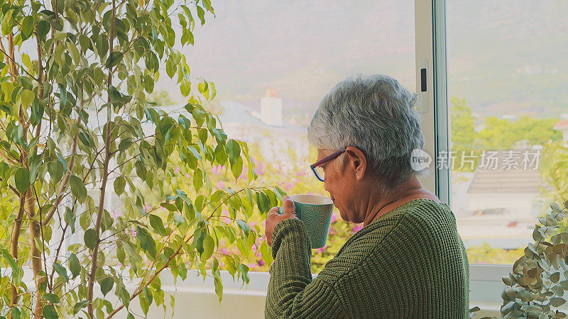 照片中，一位年长的女士一边喝着茶，一边从公寓的窗户往外看
