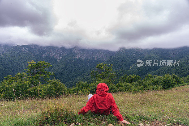 秋天的游客穿着红夹克坐在草地上看美丽的山景。热爱大自然。