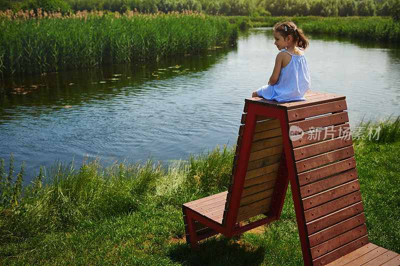 一个穿着蓝色裙子，梳着两个马尾辫的可爱女孩坐在一张木凳上，愉快地看着乡村里美丽的河的景色